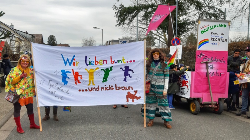 Zwei kostümierte Umzusgteilnehmerinne mit einem großen Transparent mit der Aufschirft "Wir lieben bunt, nicht braun", im Hintergrund ein Handkarren mit der Aufschrift "Liebes Deutschalnd, kannst Du mal nach den Rechten sehen?"