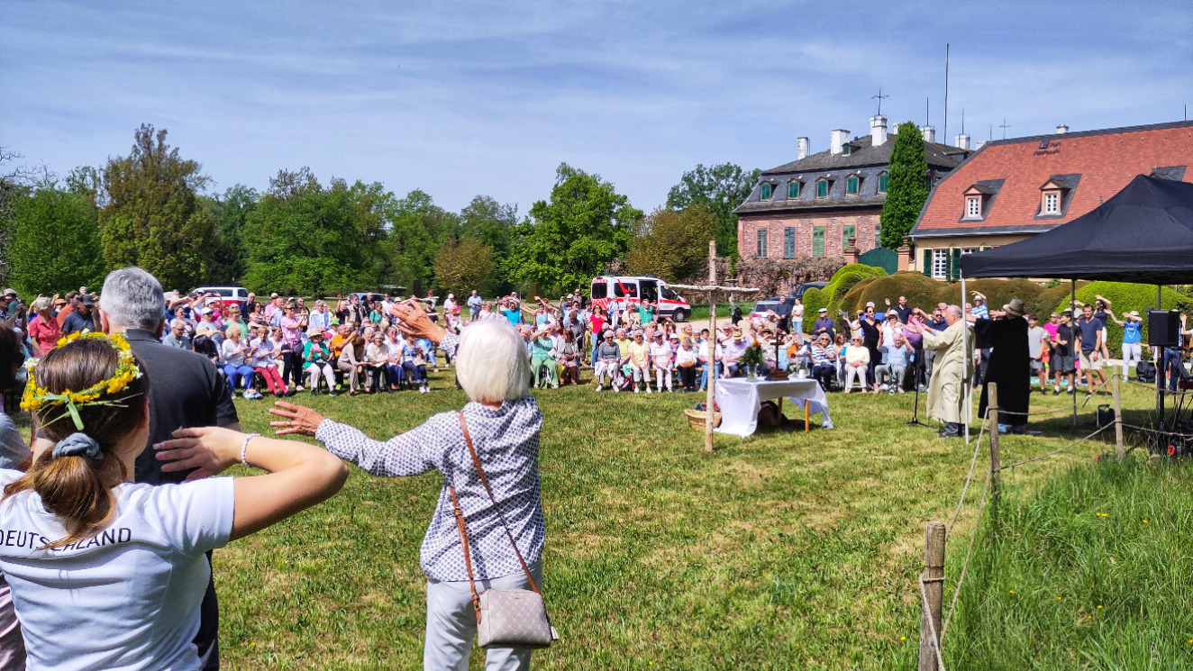 Gottesdienst im Garten von Schloss Wolfsgarten: Viele Menschen stehen auf der Wiese, die beiden Pfarrer unter einem Sonnenschutz, in der Mitte ein Kreuz aus zwei Birkenstämmen, im Vordergrund schenken Menschen ihre Arme im Takt zur Musik