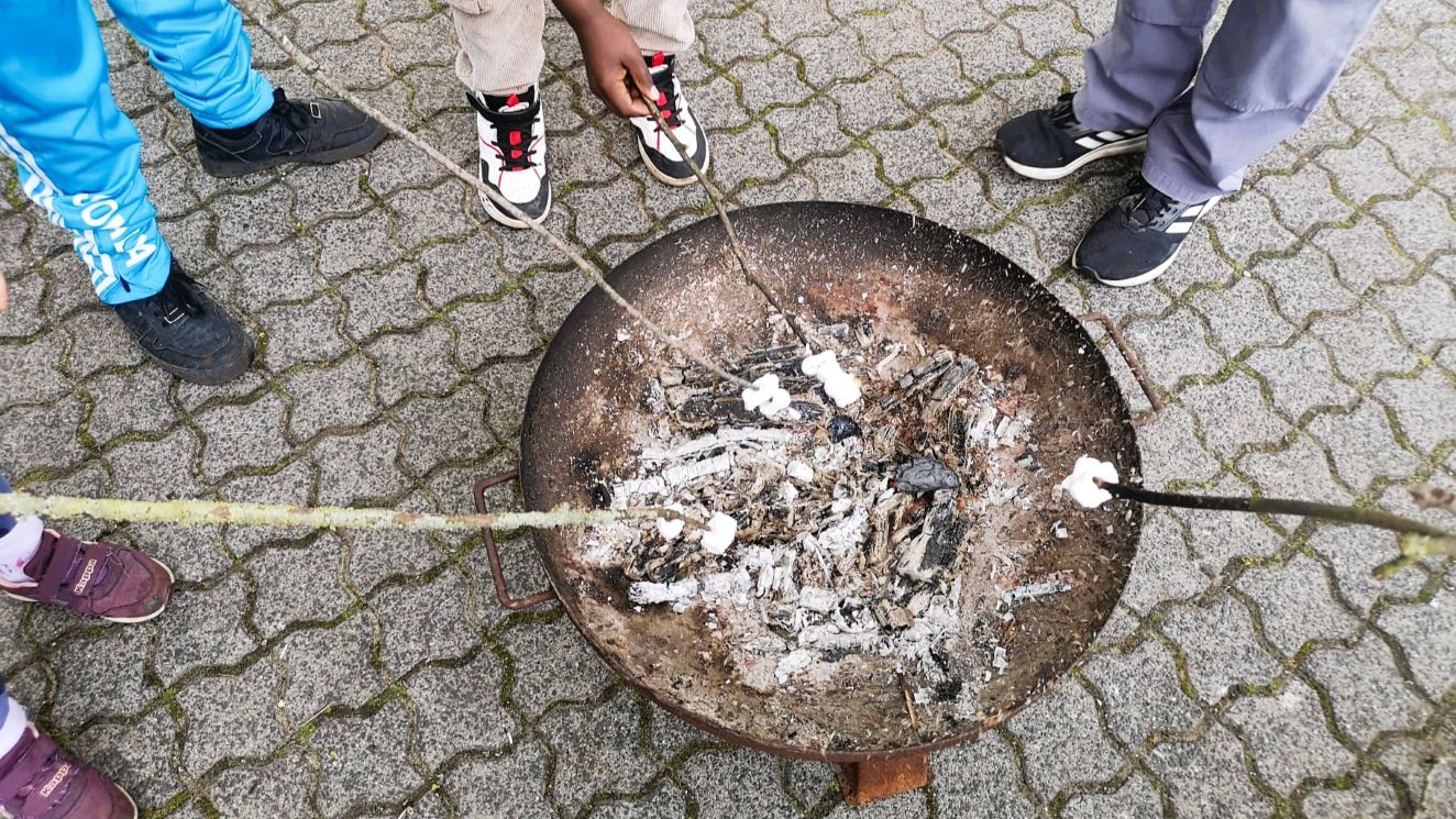 Eine Feuerschale, über der Marshmallows an Stöcken schmoren - drumherum Kinderbeine und bei einem Stzock sieht man die haltende Hand